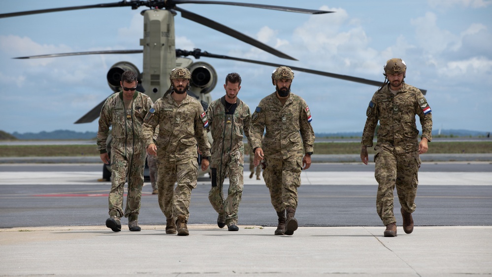 Five military personnel in camouflage uniforms walk across a tarmac or airfield away from a large tandem-rotor helicopter, identified as a CH-47 Chinook. The individuals wear various patches and insignia, with some wearing tactical helmets. The setting appears to be a joint or multinational military operation based on the variety of patches visible.