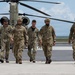 Five military personnel in camouflage uniforms walk across a tarmac or airfield away from a large tandem-rotor helicopter, identified as a CH-47 Chinook. The individuals wear various patches and insignia, with some wearing tactical helmets. The setting appears to be a joint or multinational military operation based on the variety of patches visible.