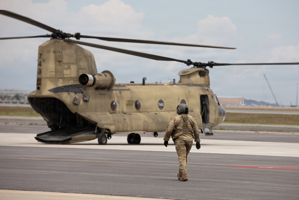 A U.S. Army soldier in full flight gear walks toward a CH-47 Chinook tandem-rotor helicopter on a tarmac. The helicopter bears the tail number 07-08743 and the number 743 on its fuselage, with 'UNITED STATES' markings visible. The scene appears to be at a military airfield with industrial structures visible in the background.