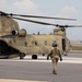 A U.S. Army soldier in full flight gear walks toward a CH-47 Chinook tandem-rotor helicopter on a tarmac. The helicopter bears the tail number 07-08743 and the number 743 on its fuselage, with 'UNITED STATES' markings visible. The scene appears to be at a military airfield with industrial structures visible in the background.
