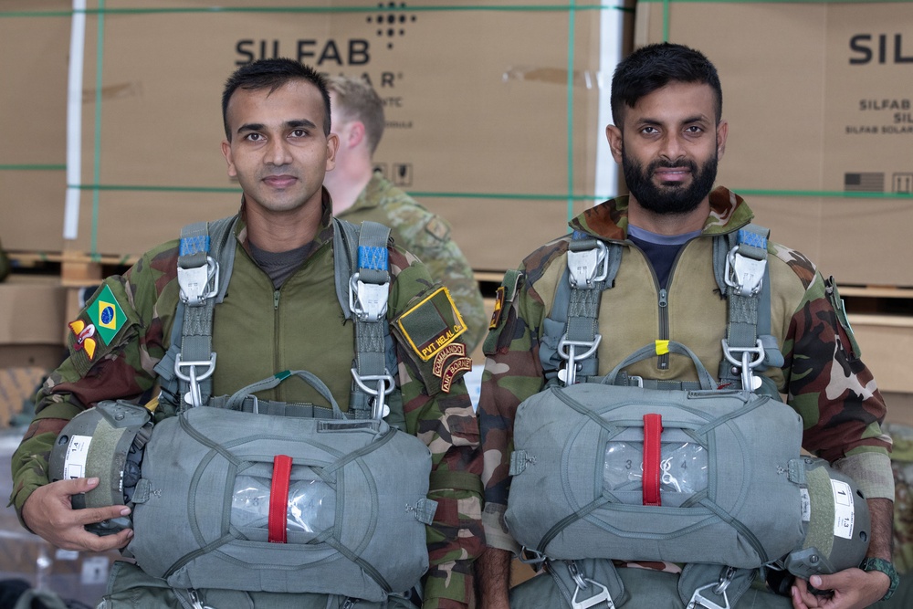 Two military personnel in camouflage uniforms are posed together wearing parachute harnesses and holding parachute packs, suggesting they are preparing for or have completed a airborne/paratrooper exercise. One individual displays a Brazilian flag patch on their left shoulder along with additional unit patches. They are standing in front of stacked 'SILFAB Solar' branded crates in what appears to be a warehouse or hangar setting.