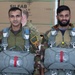 Two military personnel in camouflage uniforms are posed together wearing parachute harnesses and holding parachute packs, suggesting they are preparing for or have completed a airborne/paratrooper exercise. One individual displays a Brazilian flag patch on their left shoulder along with additional unit patches. They are standing in front of stacked 'SILFAB Solar' branded crates in what appears to be a warehouse or hangar setting.