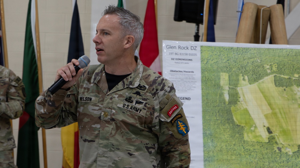A U.S. Army soldier named Nelson is speaking into a microphone during what appears to be a military briefing or presentation. He is standing in front of a map board labeled 'Glen Rock DZ' (Drop Zone) with aerial imagery and grid coordinates visible. Multiple national flags are displayed in the background, suggesting a multinational military event.