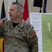 A U.S. Army soldier named Nelson is speaking into a microphone during what appears to be a military briefing or presentation. He is standing in front of a map board labeled 'Glen Rock DZ' (Drop Zone) with aerial imagery and grid coordinates visible. Multiple national flags are displayed in the background, suggesting a multinational military event.