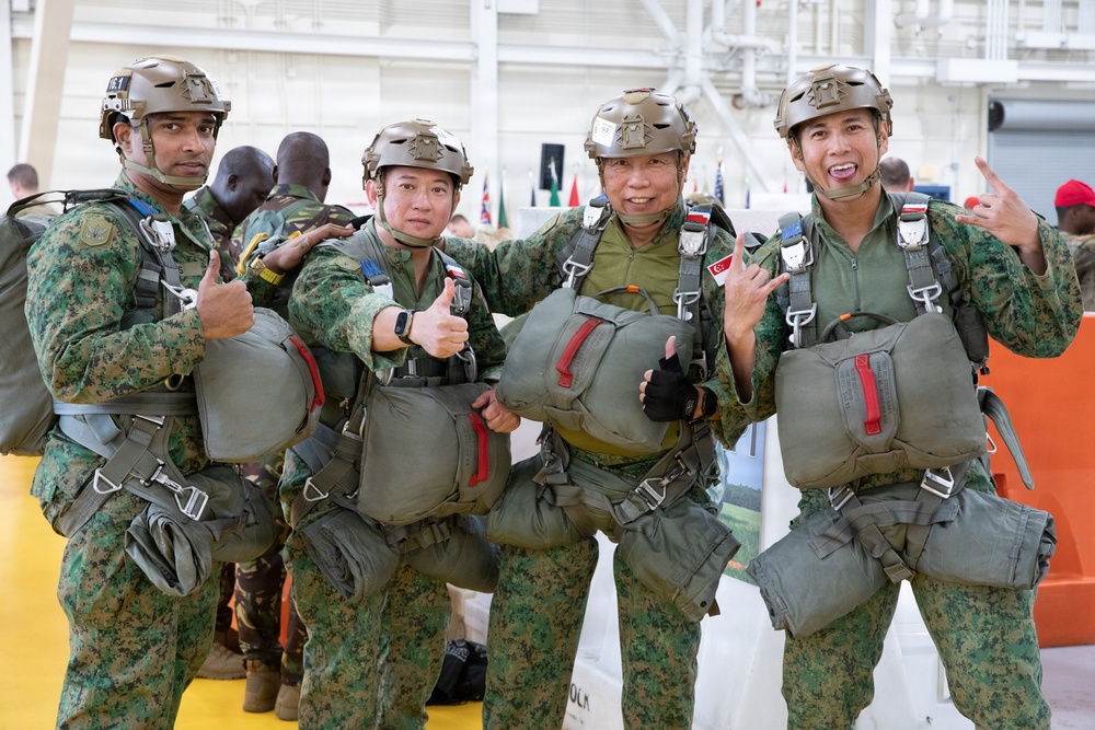 Four military personnel in matching camouflage uniforms pose together for a photo, appearing cheerful and making thumbs-up and 'shaka' hand gestures. They are fully equipped with parachute harnesses, reserve parachute packs, and tactical helmets, suggesting they are preparing for or have completed a parachute jump. The setting appears to be an indoor military facility or hangar with international flags visible in the background.