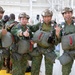 Four military personnel in matching camouflage uniforms pose together for a photo, appearing cheerful and making thumbs-up and 'shaka' hand gestures. They are fully equipped with parachute harnesses, reserve parachute packs, and tactical helmets, suggesting they are preparing for or have completed a parachute jump. The setting appears to be an indoor military facility or hangar with international flags visible in the background.