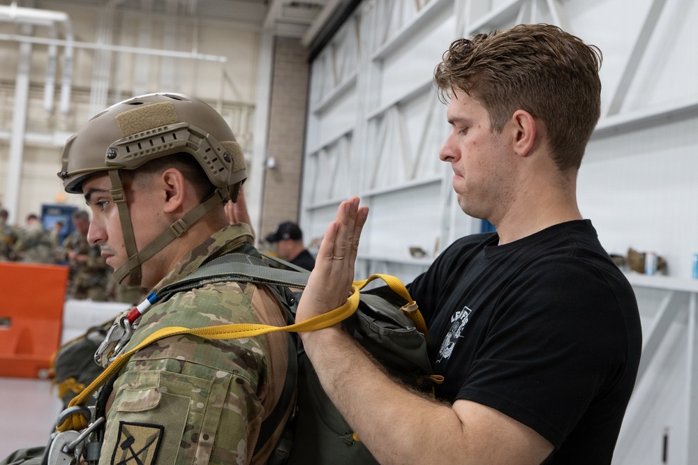 A man in a black t-shirt is inspecting or adjusting parachute equipment on a soldier wearing multicam military uniform and a tactical helmet. The activity appears to be a pre-jump equipment check taking place inside a military hangar. Additional personnel in military uniforms are visible in the background.