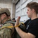 A man in a black t-shirt is inspecting or adjusting parachute equipment on a soldier wearing multicam military uniform and a tactical helmet. The activity appears to be a pre-jump equipment check taking place inside a military hangar. Additional personnel in military uniforms are visible in the background.