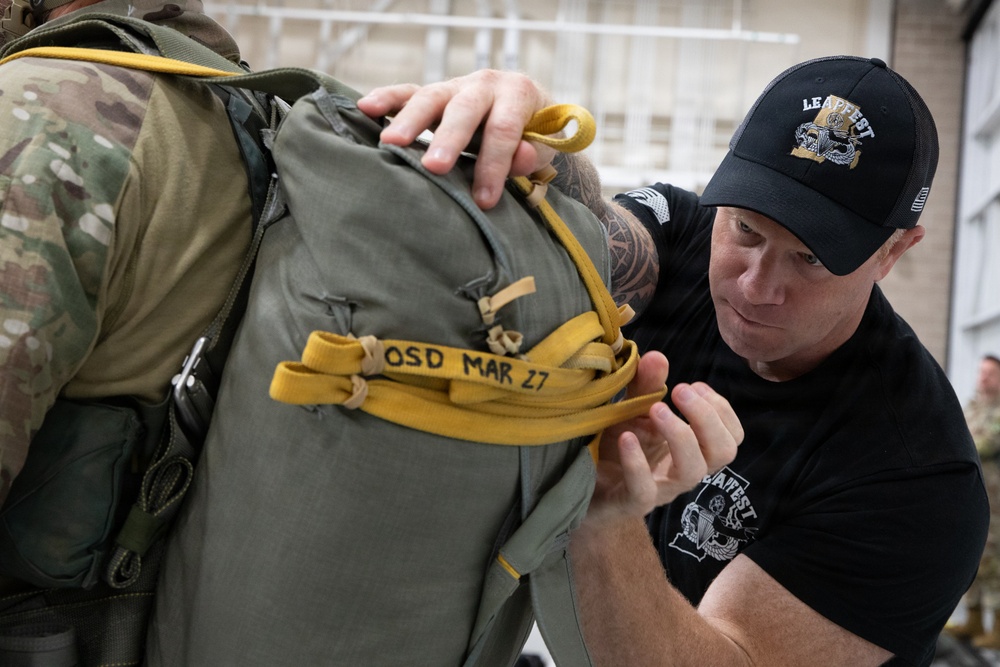 A man wearing a black 'LEAPFEST' cap and t-shirt is inspecting or adjusting a military parachute pack being worn by a soldier in camouflage uniform. The parachute pack has yellow straps with the marking 'OSD MAR 27' written on one of the straps. This appears to be a pre-jump equipment inspection at a military airborne training event.