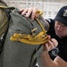 A man wearing a black 'LEAPFEST' cap and t-shirt is inspecting or adjusting a military parachute pack being worn by a soldier in camouflage uniform. The parachute pack has yellow straps with the marking 'OSD MAR 27' written on one of the straps. This appears to be a pre-jump equipment inspection at a military airborne training event.