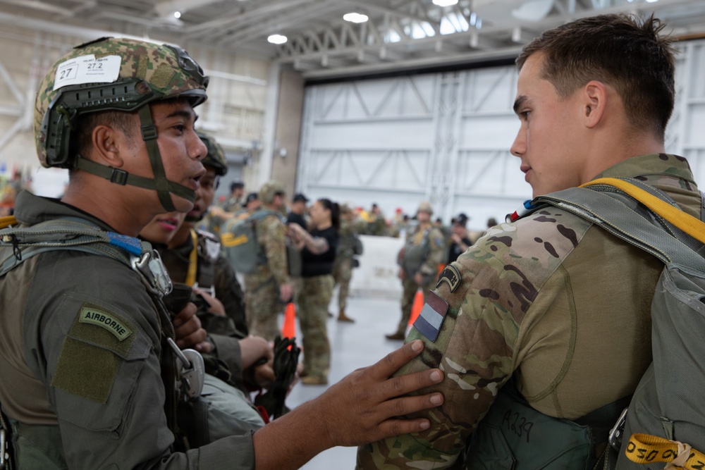 Two military personnel in a hangar are conducting a pre-jump equipment check, with one soldier wearing an Airborne tab and a helmet numbered 27/27.2 inspecting the parachute harness of another soldier wearing a Netherlands flag patch. The setting appears to be a multinational airborne operation or exercise, with numerous other uniformed personnel visible in the background. The environment is a large military aircraft hangar with high ceilings and industrial lighting.