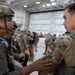 Two military personnel in a hangar are conducting a pre-jump equipment check, with one soldier wearing an Airborne tab and a helmet numbered 27/27.2 inspecting the parachute harness of another soldier wearing a Netherlands flag patch. The setting appears to be a multinational airborne operation or exercise, with numerous other uniformed personnel visible in the background. The environment is a large military aircraft hangar with high ceilings and industrial lighting.
