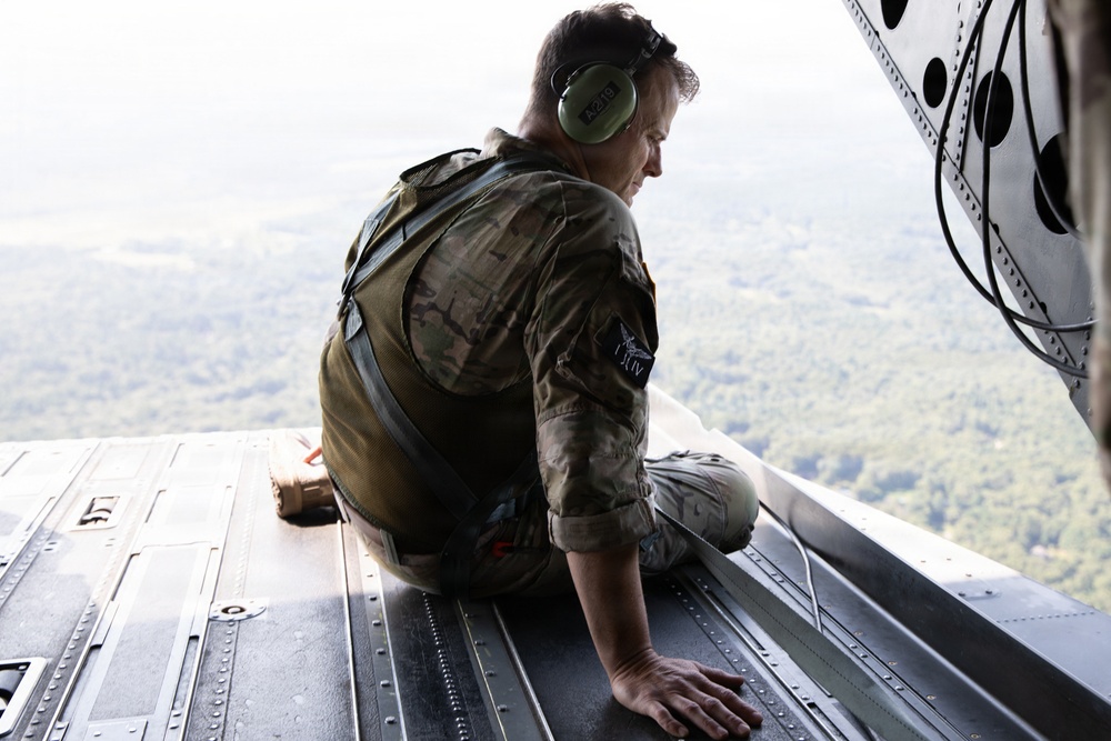 A military personnel member in camouflage uniform is seated on the open rear ramp of a military aircraft in flight, looking down at the terrain below. The individual is wearing green ear protection headphones and a tactical vest, positioned at the edge of the open cargo ramp. The landscape visible below appears to be forested or vegetated terrain viewed from a significant altitude.