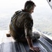 A military personnel member in camouflage uniform is seated on the open rear ramp of a military aircraft in flight, looking down at the terrain below. The individual is wearing green ear protection headphones and a tactical vest, positioned at the edge of the open cargo ramp. The landscape visible below appears to be forested or vegetated terrain viewed from a significant altitude.