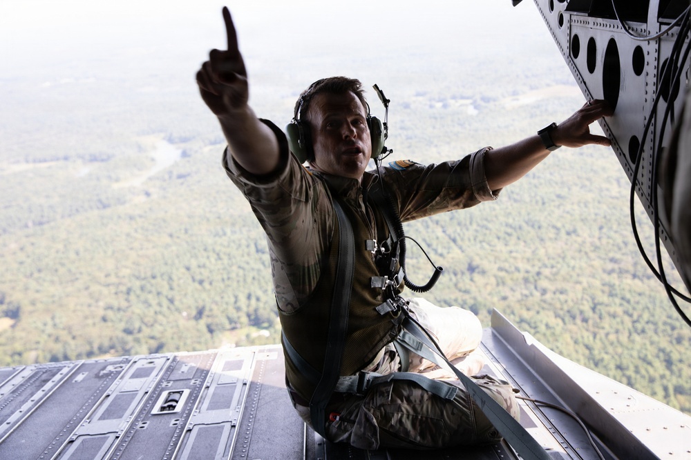 A military aircrew member is seated at the open rear ramp of a large military transport aircraft in flight, pointing with one finger while steadying himself against the aircraft structure. The individual is wearing flight gear including a helmet with communications headset, harness/safety straps, and olive drab flight suit. Below the aircraft, a densely forested landscape with a visible river or waterway is seen from significant altitude.