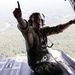 A military aircrew member is seated at the open rear ramp of a large military transport aircraft in flight, pointing with one finger while steadying himself against the aircraft structure. The individual is wearing flight gear including a helmet with communications headset, harness/safety straps, and olive drab flight suit. Below the aircraft, a densely forested landscape with a visible river or waterway is seen from significant altitude.