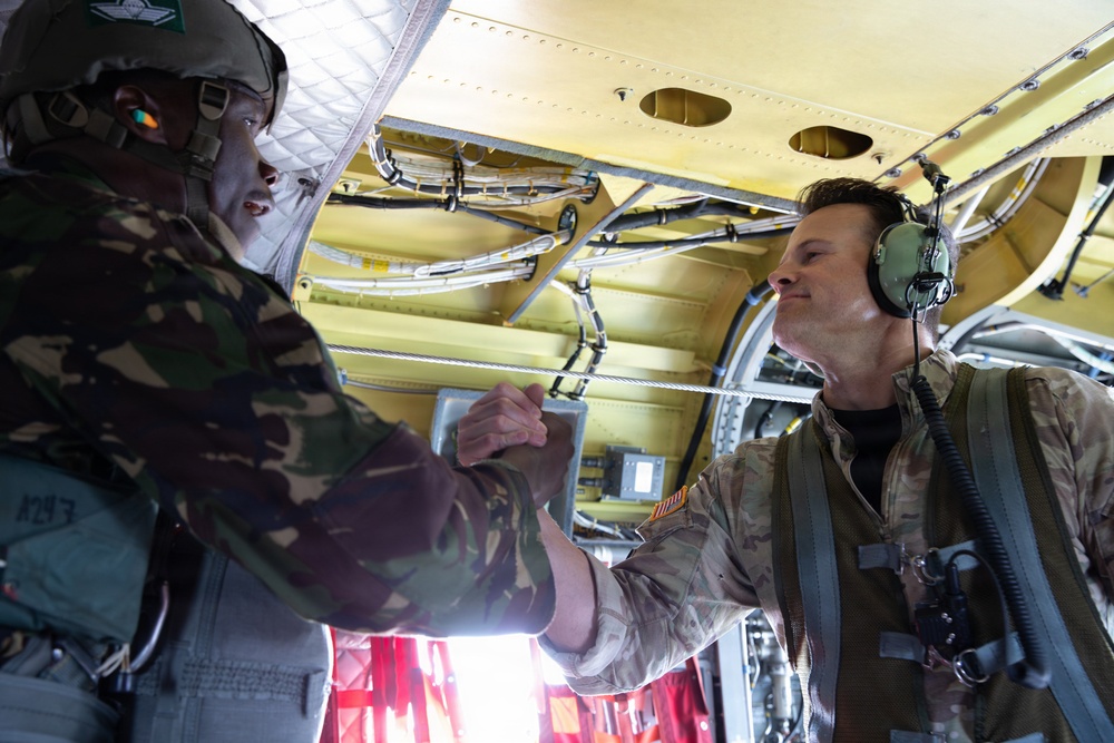 Two military personnel are inside what appears to be a military transport aircraft, engaging in a fist bump or handshake gesture. One individual on the left wears a camouflage uniform with a helmet, while the person on the right wears a flight suit with aviation headset/headphones. The interior of the aircraft is visible with yellow-painted structural components, cables, and rigging.