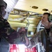 Two military personnel are inside what appears to be a military transport aircraft, engaging in a fist bump or handshake gesture. One individual on the left wears a camouflage uniform with a helmet, while the person on the right wears a flight suit with aviation headset/headphones. The interior of the aircraft is visible with yellow-painted structural components, cables, and rigging.