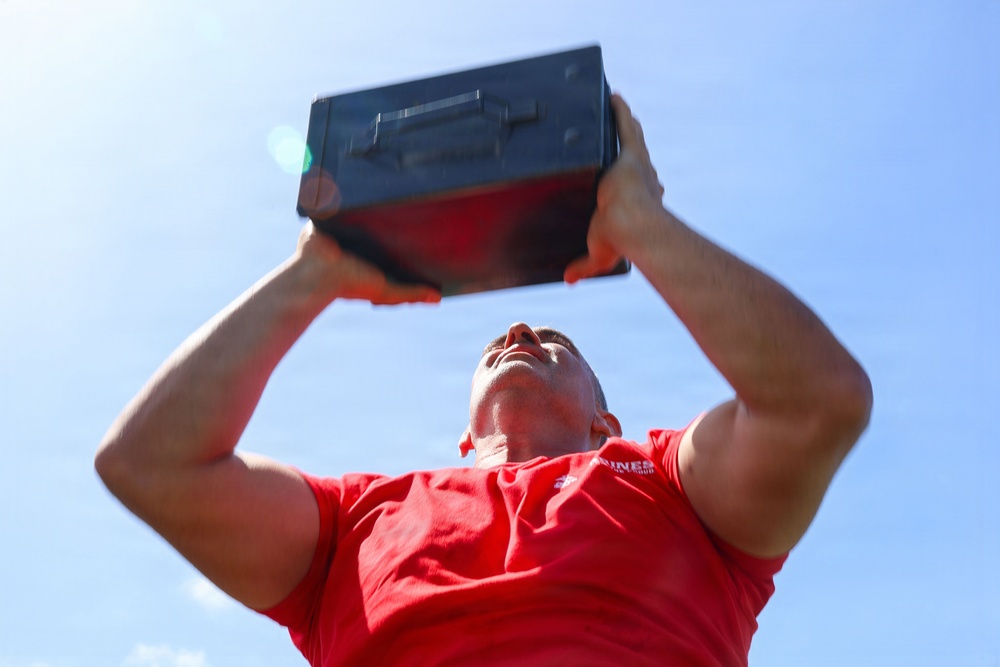 A muscular man wearing a red t-shirt is photographed from a low angle while holding a dark metal ammo can overhead with both arms extended. The shot is taken from below looking upward, creating a dramatic perspective against a bright blue sky. The activity appears to be a military-style fitness exercise involving overhead lifts with an ammunition can.
