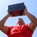 A muscular man wearing a red t-shirt is photographed from a low angle while holding a dark metal ammo can overhead with both arms extended. The shot is taken from below looking upward, creating a dramatic perspective against a bright blue sky. The activity appears to be a military-style fitness exercise involving overhead lifts with an ammunition can.