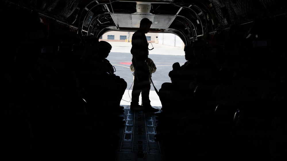 The image shows the interior of a military transport aircraft with the rear cargo ramp open, looking out onto a tarmac or airfield. Several personnel are visible in silhouette, with one individual standing at the open ramp and others seated along the sides of the aircraft. The strong backlight from outside creates a high-contrast silhouette effect, obscuring individual details.