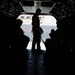 The image shows the interior of a military transport aircraft with the rear cargo ramp open, looking out onto a tarmac or airfield. Several personnel are visible in silhouette, with one individual standing at the open ramp and others seated along the sides of the aircraft. The strong backlight from outside creates a high-contrast silhouette effect, obscuring individual details.