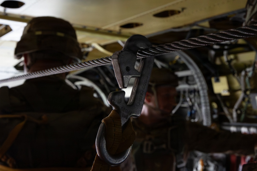 The image shows a close-up of a military static line snap hook attached to a steel anchor cable inside what appears to be a military transport aircraft. Two soldiers in camouflage uniforms are visible in the background, out of focus. The setting suggests airborne operations preparation or training.