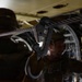 The image shows a close-up of a military static line snap hook attached to a steel anchor cable inside what appears to be a military transport aircraft. Two soldiers in camouflage uniforms are visible in the background, out of focus. The setting suggests airborne operations preparation or training.