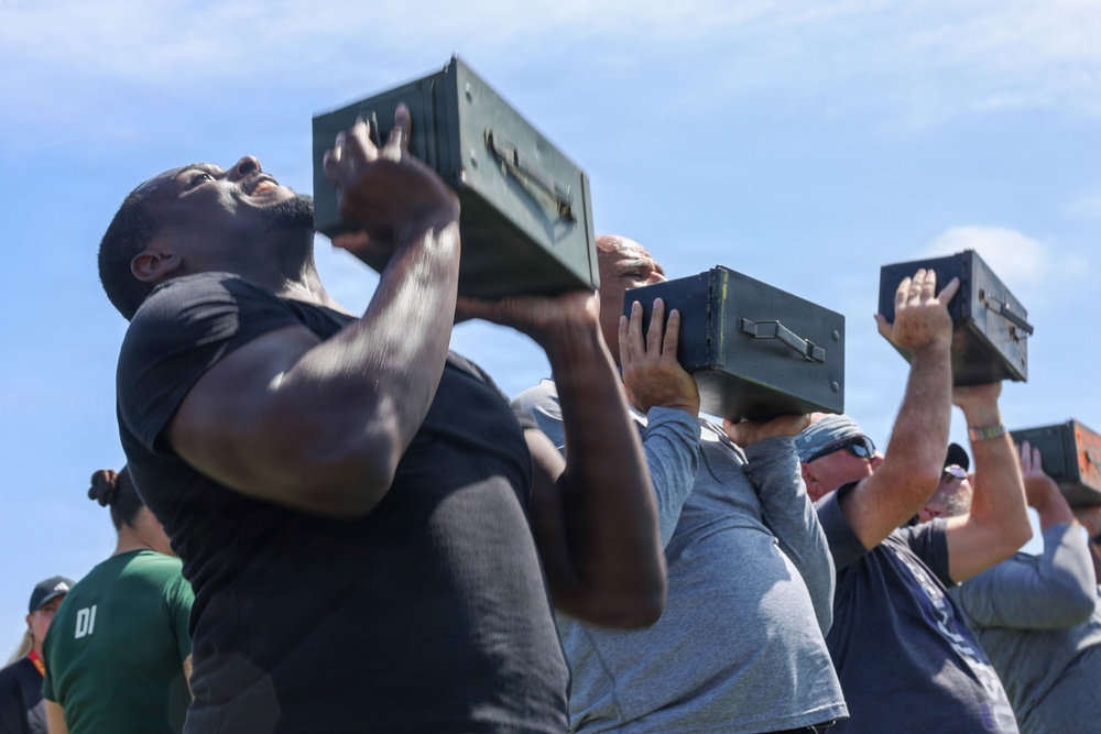 A group of individuals are lifting military-style ammunition cans overhead in what appears to be a physical fitness or training exercise. The foreground features a muscular man in a black t-shirt straining to press the metal box overhead, with several other participants visible behind him performing the same movement. The setting appears to be an outdoor area under a partly cloudy sky.