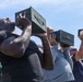 A group of individuals are lifting military-style ammunition cans overhead in what appears to be a physical fitness or training exercise. The foreground features a muscular man in a black t-shirt straining to press the metal box overhead, with several other participants visible behind him performing the same movement. The setting appears to be an outdoor area under a partly cloudy sky.