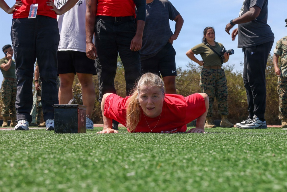 A woman in a red Marines t-shirt is performing a push-up or low crawl exercise on an artificial turf field. She is surrounded by a group of observers, including individuals in civilian clothing and at least two people in U.S. Marine Corps camouflage uniforms. The setting appears to be a military fitness demonstration or recruiting event.