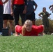 A woman in a red Marines t-shirt is performing a push-up or low crawl exercise on an artificial turf field. She is surrounded by a group of observers, including individuals in civilian clothing and at least two people in U.S. Marine Corps camouflage uniforms. The setting appears to be a military fitness demonstration or recruiting event.