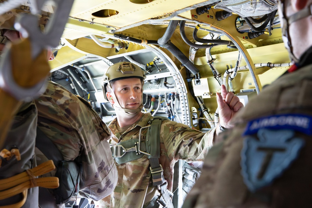 Military personnel are inside what appears to be a military aircraft, likely during a pre-jump briefing or inspection. At least three soldiers in camouflage uniforms and tactical gear are visible, with one prominently displaying an 'Airborne' patch on their shoulder. The interior of the aircraft shows exposed wiring, hydraulic lines, and structural components painted in yellow-green primer.