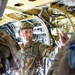 Military personnel are inside what appears to be a military aircraft, likely during a pre-jump briefing or inspection. At least three soldiers in camouflage uniforms and tactical gear are visible, with one prominently displaying an 'Airborne' patch on their shoulder. The interior of the aircraft shows exposed wiring, hydraulic lines, and structural components painted in yellow-green primer.