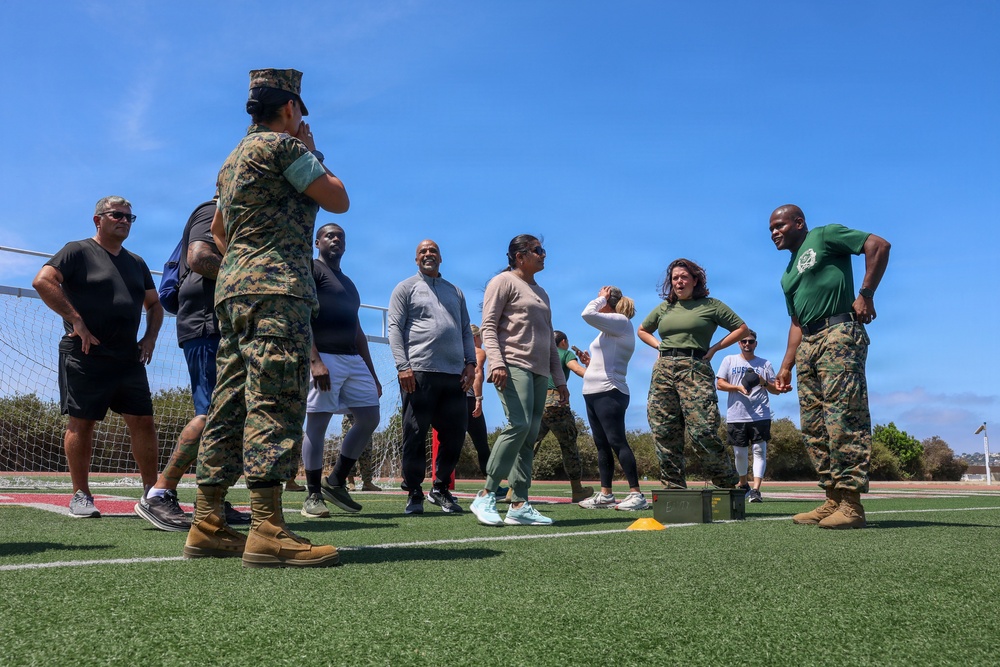 A group of civilians and military personnel are gathered on an outdoor athletic field with artificial turf. Two Marines in camouflage uniforms appear to be instructing or leading a group of civilians in what appears to be a physical training or orientation event. The setting includes a soccer goal post and yellow training cones, suggesting an organized fitness or community engagement activity.