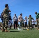 A group of civilians and military personnel are gathered on an outdoor athletic field with artificial turf. Two Marines in camouflage uniforms appear to be instructing or leading a group of civilians in what appears to be a physical training or orientation event. The setting includes a soccer goal post and yellow training cones, suggesting an organized fitness or community engagement activity.