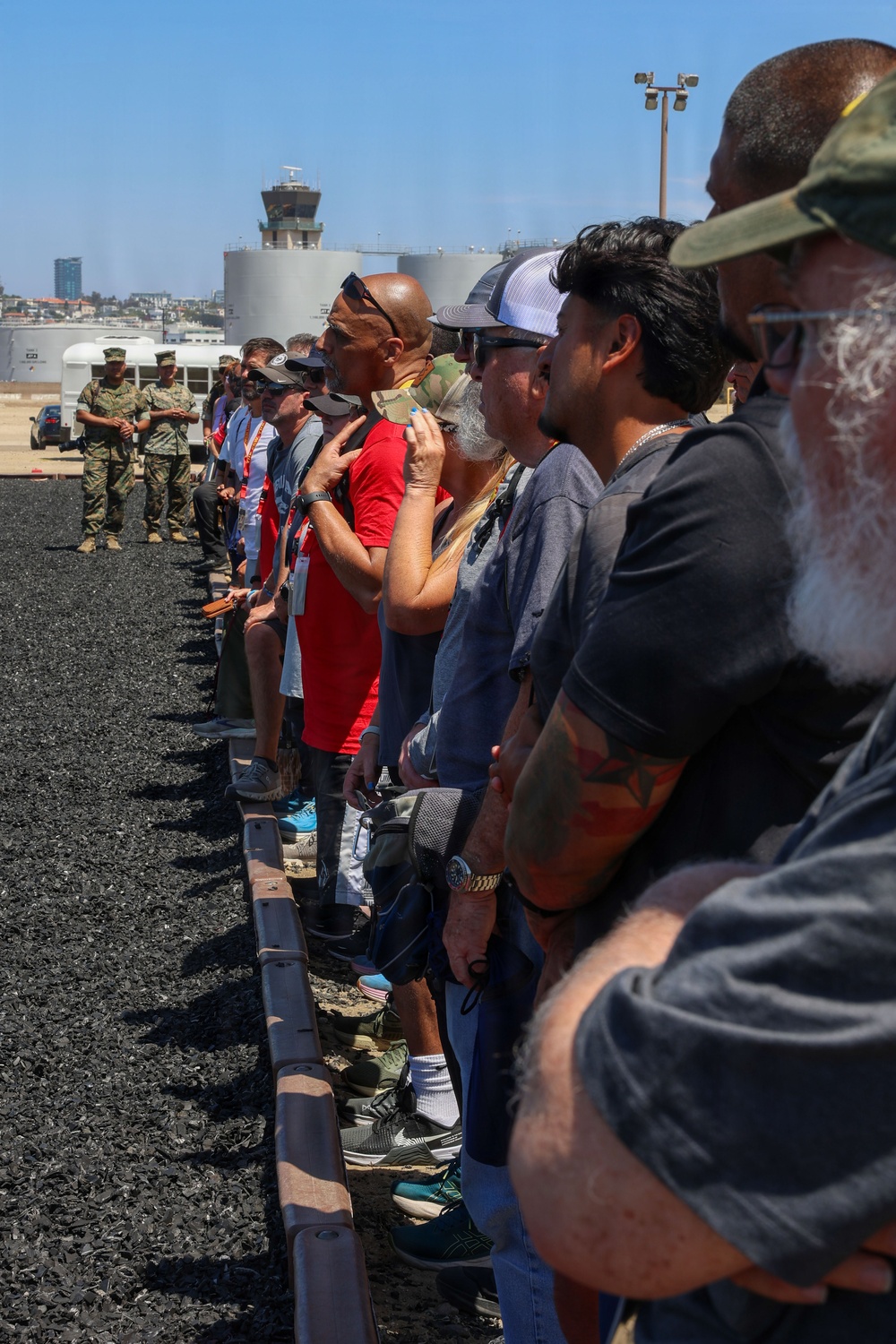 A group of civilians stands in a line on a tarmac or airfield surface, appearing to observe something off-camera to their right. In the background, two uniformed military personnel (appearing to be U.S. Marines in camouflage uniforms) stand near a white bus, suggesting this is an organized tour or demonstration event at a military installation. An air traffic control tower and industrial storage tanks are visible in the background, consistent with a military airfield setting.