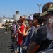 A group of civilians stands in a line on a tarmac or airfield surface, appearing to observe something off-camera to their right. In the background, two uniformed military personnel (appearing to be U.S. Marines in camouflage uniforms) stand near a white bus, suggesting this is an organized tour or demonstration event at a military installation. An air traffic control tower and industrial storage tanks are visible in the background, consistent with a military airfield setting.