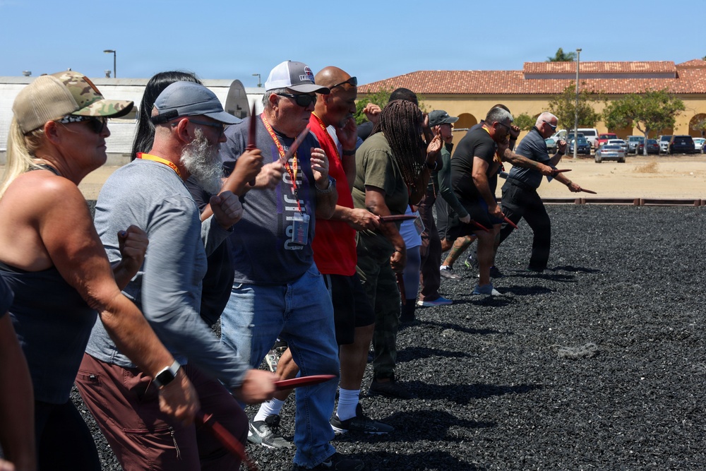 A group of adults are participating in an outdoor training or self-defense class, holding what appear to be training sticks or batons in a coordinated stance. The activity takes place on a dark gravel surface in front of a building with a red-tiled roof, suggesting a military base or institutional facility. Participants are wearing casual civilian clothing and some have lanyards, indicating a structured program or event.