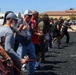 A group of adults are participating in an outdoor training or self-defense class, holding what appear to be training sticks or batons in a coordinated stance. The activity takes place on a dark gravel surface in front of a building with a red-tiled roof, suggesting a military base or institutional facility. Participants are wearing casual civilian clothing and some have lanyards, indicating a structured program or event.