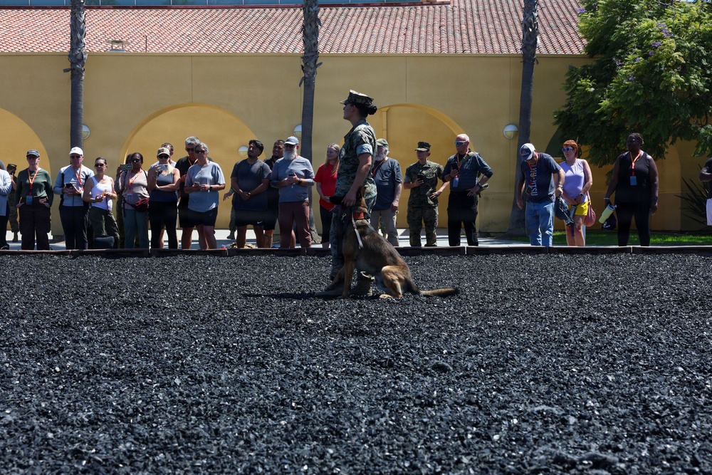 A military handler in camouflage uniform and cover (cap) stands on a dark gravel surface with a military working dog (appears to be a Belgian Malinois) during what appears to be a public demonstration. A crowd of civilian spectators and a few uniformed personnel observe from behind a boundary line in front of a yellow stucco building with arched architectural details. The setting appears to be a military base open house or community relations event.