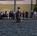 A military handler in camouflage uniform and cover (cap) stands on a dark gravel surface with a military working dog (appears to be a Belgian Malinois) during what appears to be a public demonstration. A crowd of civilian spectators and a few uniformed personnel observe from behind a boundary line in front of a yellow stucco building with arched architectural details. The setting appears to be a military base open house or community relations event.
