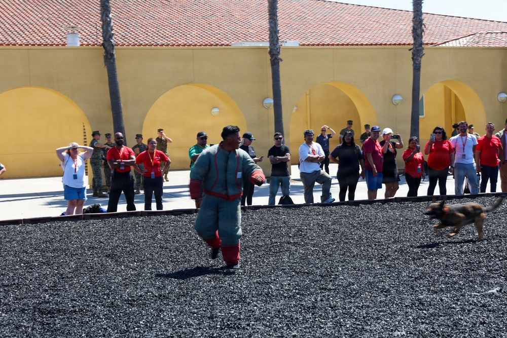A military working dog demonstration is taking place at what appears to be a military installation, likely a Marine Corps base based on the visible camouflage uniforms. A decoy wearing a padded bite suit is being pursued by a German Shepherd across a gravel training area while a crowd of spectators, many wearing red lanyards, observe from the perimeter. The yellow stucco building with arched architecture and palm trees in the background is consistent with a Southern California military base.