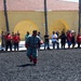 A military working dog demonstration is taking place at what appears to be a military installation, likely a Marine Corps base based on the visible camouflage uniforms. A decoy wearing a padded bite suit is being pursued by a German Shepherd across a gravel training area while a crowd of spectators, many wearing red lanyards, observe from the perimeter. The yellow stucco building with arched architecture and palm trees in the background is consistent with a Southern California military base.