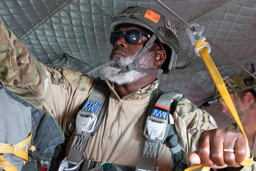 A military paratrooper is inside an aircraft, preparing for a jump operation. The individual is wearing full parachute harness equipment, a tactical helmet marked with the number '42', and is holding a yellow static line. A second person in military uniform is partially visible in the background.
