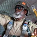 A military paratrooper is inside an aircraft, preparing for a jump operation. The individual is wearing full parachute harness equipment, a tactical helmet marked with the number '42', and is holding a yellow static line. A second person in military uniform is partially visible in the background.