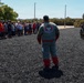 A military K-9 unit demonstration is taking place at what appears to be MCRD (Marine Corps Recruit Depot) based on the patch visible on the bite suit. A person wearing a padded protection suit with 'MCRD K-9' insignia stands in a rubber mulch training area facing a crowd of civilian spectators, while a handler with a military working dog is visible on the right side. The event appears to be a public demonstration or family day showcasing military working dog capabilities.