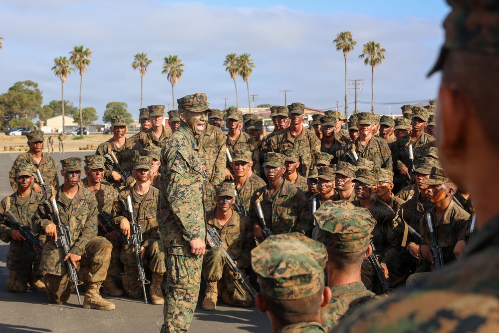 A large group of U.S. Marines in MARPAT woodland camouflage uniforms are assembled outdoors, many wearing camouflage face paint and carrying M16/M4-style rifles with blank firing adapters. A central figure, likely a senior NCO or officer, stands addressing the assembled group. The setting appears to be a military installation with palm trees and low buildings visible in the background, consistent with a Southern California base such as Camp Pendleton.