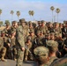 A large group of U.S. Marines in MARPAT woodland camouflage uniforms are assembled outdoors, many wearing camouflage face paint and carrying M16/M4-style rifles with blank firing adapters. A central figure, likely a senior NCO or officer, stands addressing the assembled group. The setting appears to be a military installation with palm trees and low buildings visible in the background, consistent with a Southern California base such as Camp Pendleton.