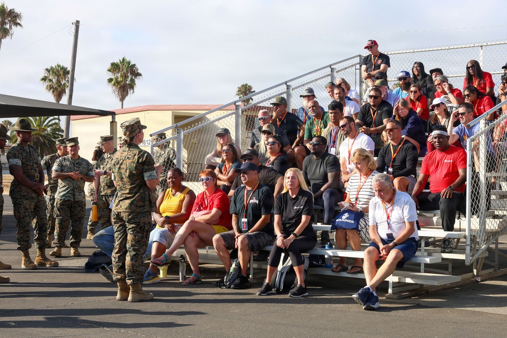 A group of U.S. Marines in camouflage uniforms are addressing a large civilian audience seated on bleachers at what appears to be a military base. The civilians, many wearing lanyards and red shirts, appear to be attending an organized event such as a family day, community outreach, or recruit graduation. Palm trees and military-style buildings are visible in the background, suggesting a Southern California installation.